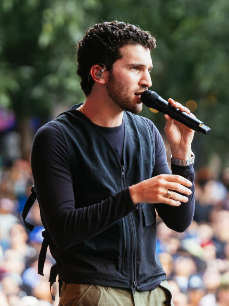 Male singer performing outdoors into a handheld microphone, wearing a black zippered jacket and watch, with a blurred crowd behind him.] , but need proper punctuation