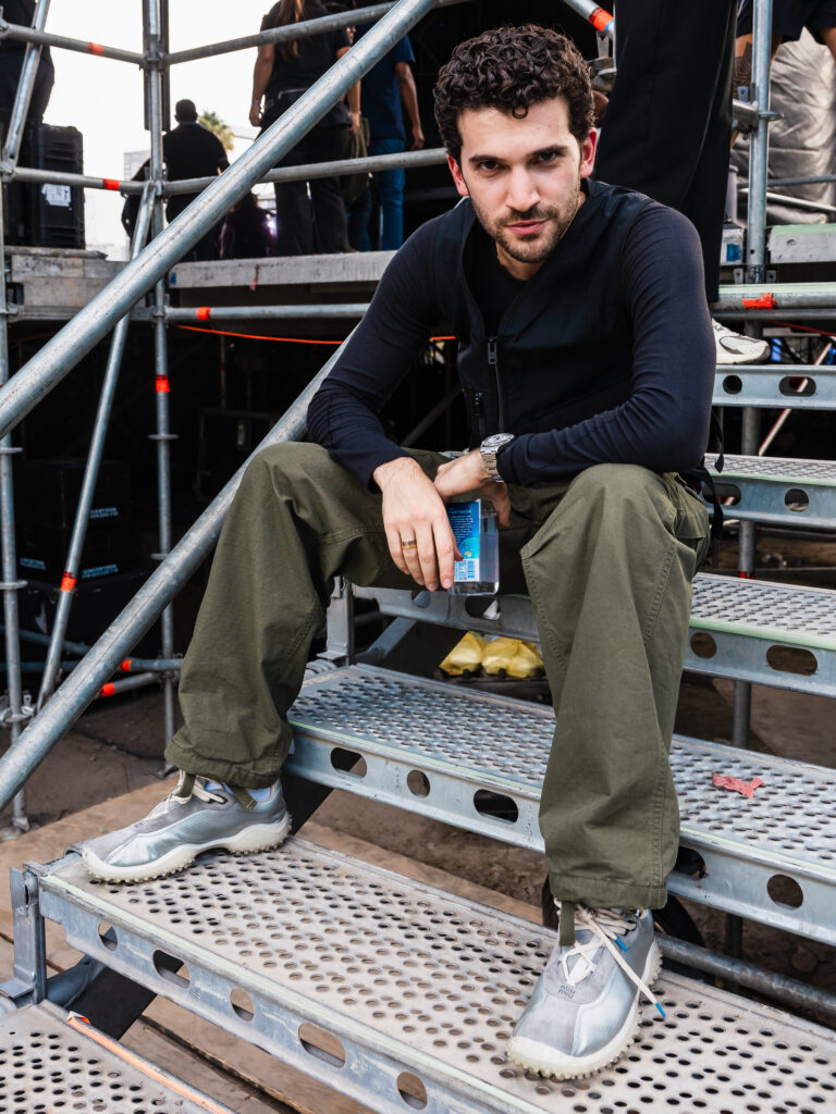 Man with curly hair sits on metal scaffold steps at an outdoor event, wearing a black long-sleeve shirt, olive pants, gray sneakers, and holding a water bottle.