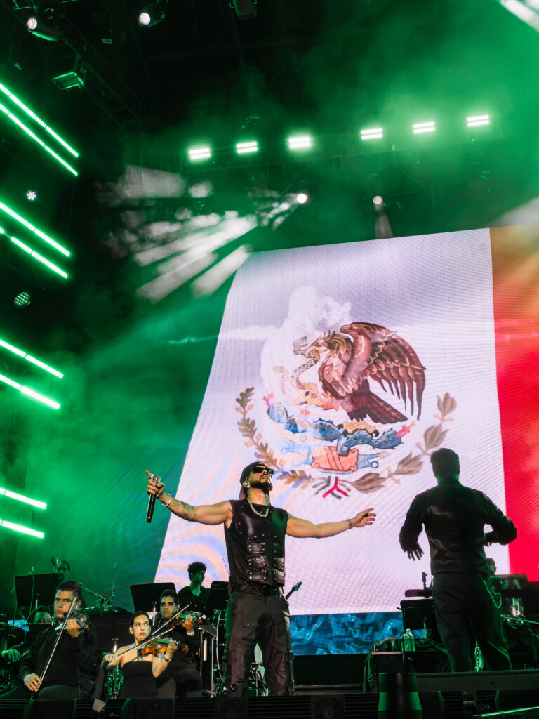 Male singer on stage with arms outstretched, a large Mexican flag with the coat of arms on a screen behind, and green stage lights for effect.