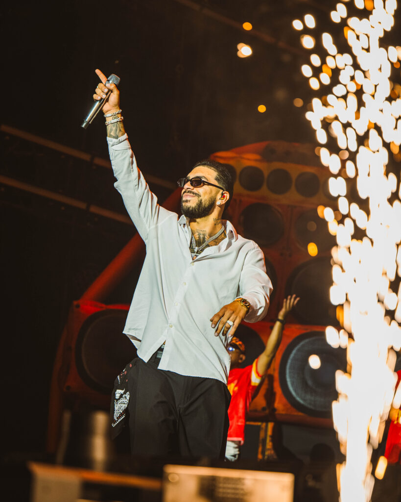 Male singer on stage raises a microphone, wearing sunglasses and a light shirt, with tattoos and bracelets visible against bright bokeh lights.